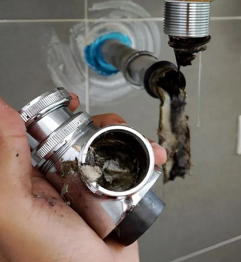 Hand holding a clogged chrome sink drain pipe under a bathroom sink.