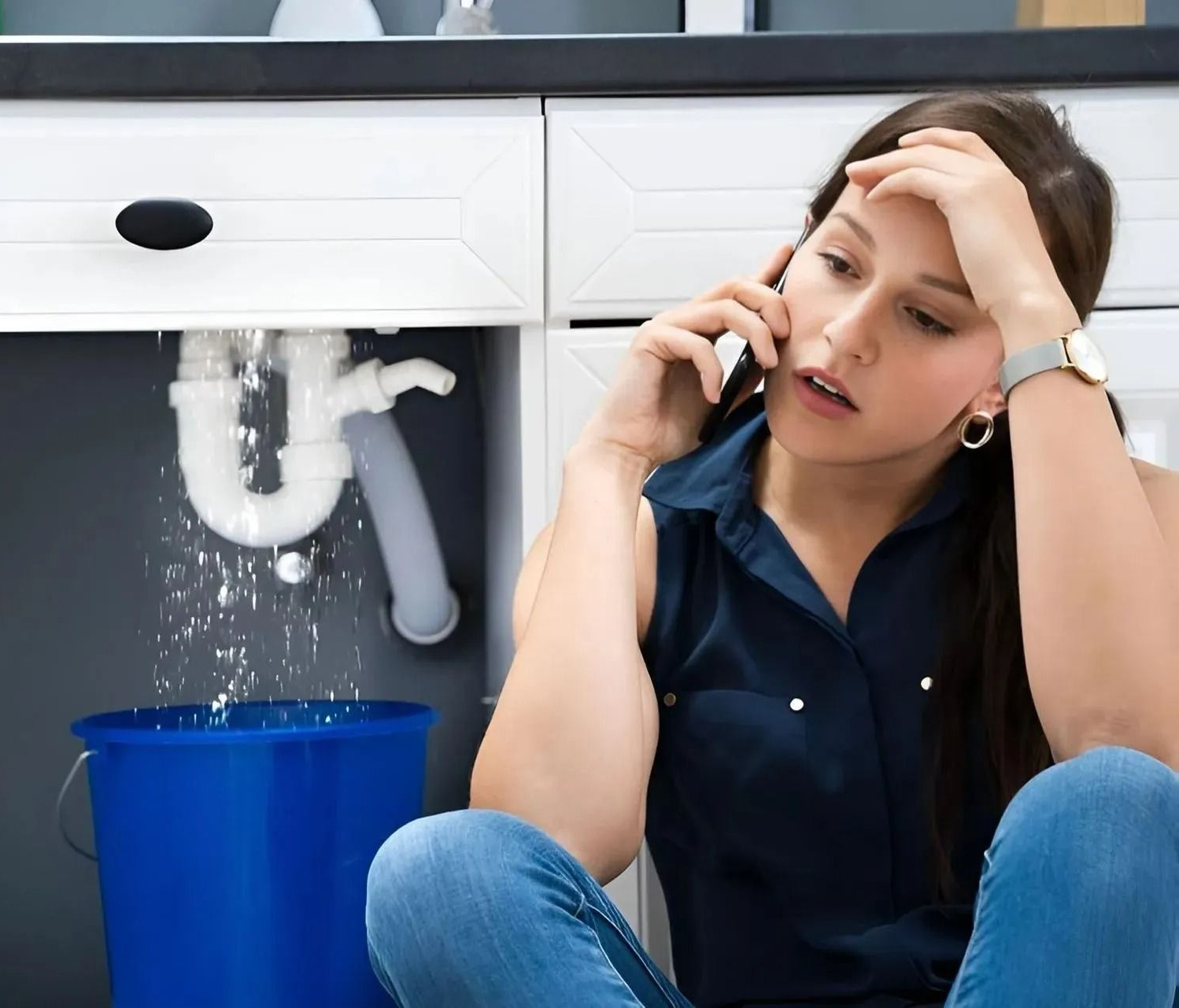 Woman on floor beside leaking sink pipe, holding phone and head, with a blue bucket catching water