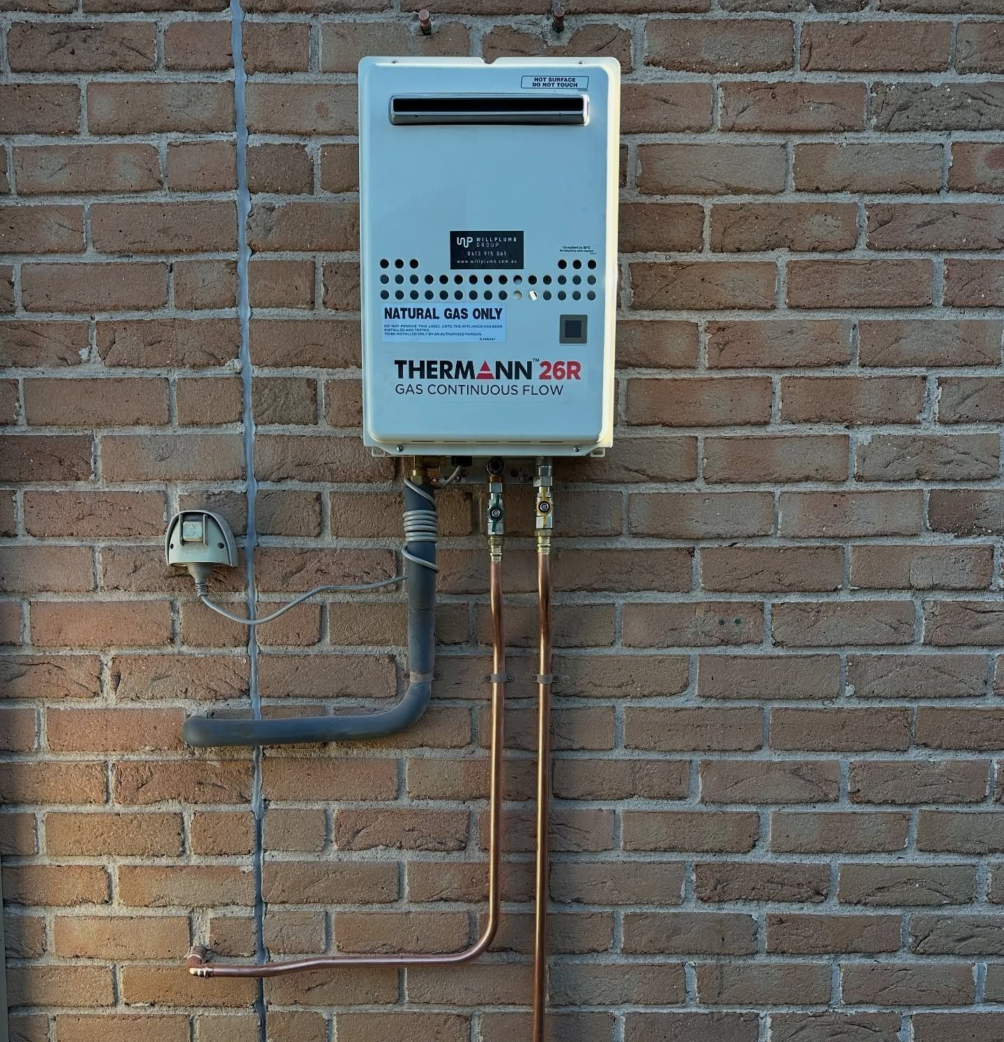 Wall-mounted gas water heater on a brick exterior wall with exposed pipes and a small meter.