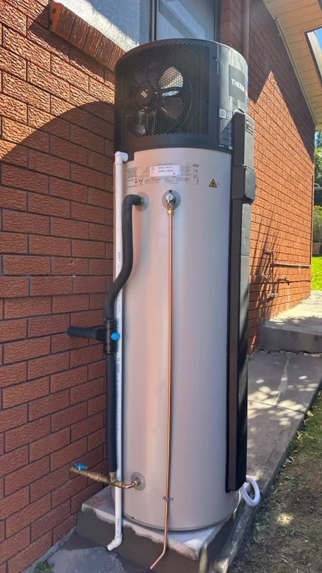 Tall silver cylindrical outdoor shower or water tank against a brick wall, with a black top and hose attachments