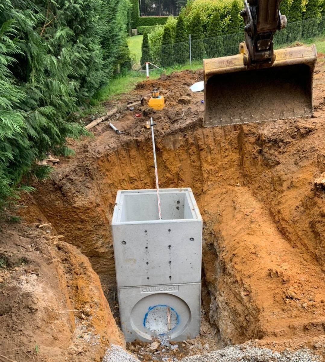 Excavator lowering a concrete utility chamber into a deep dirt pit in a backyard.