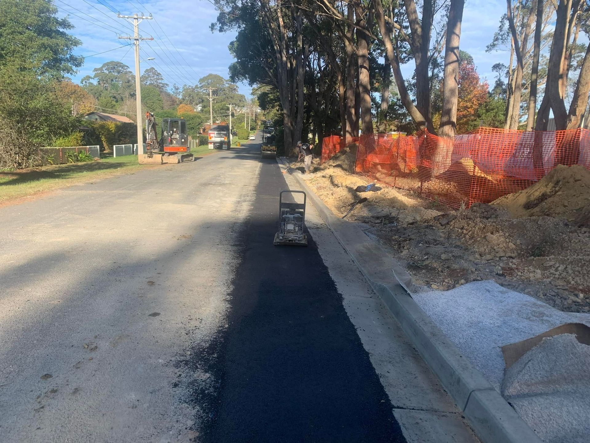 Roadwork on a sunny residential street with orange safety fencing and a newly paved dark lane.