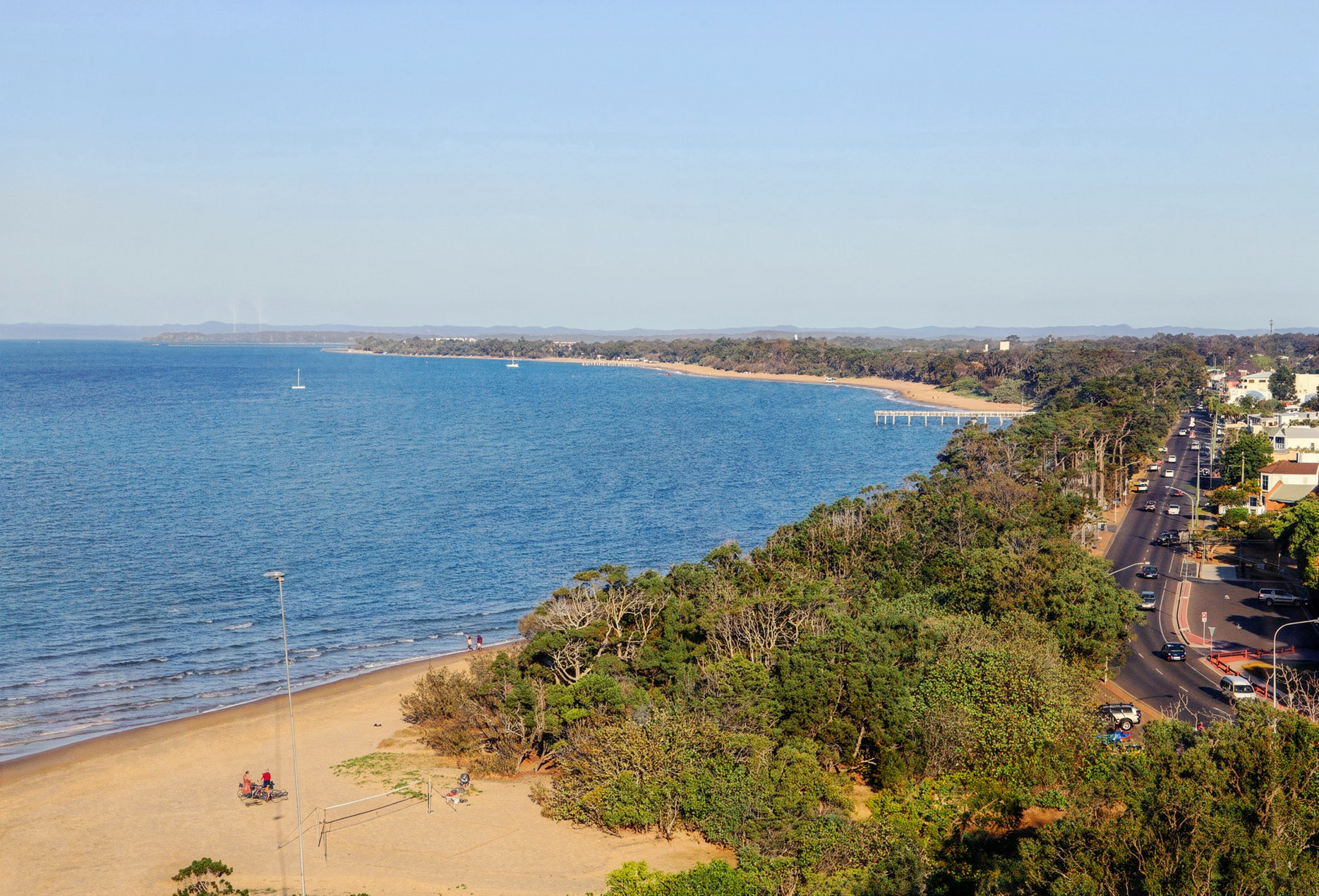 Aerial view of a sandy beach and blue coastline beside a road lined with trees and buildings