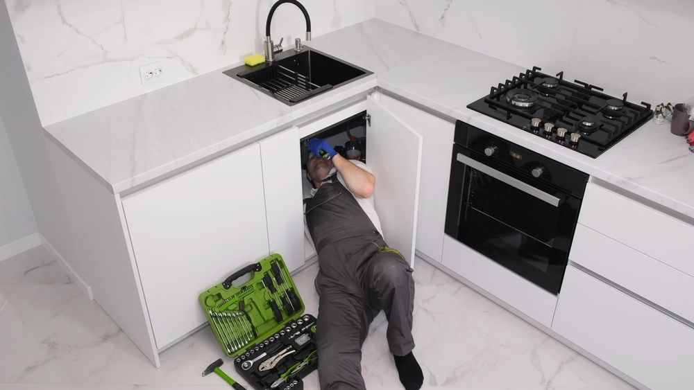 Person lying on kitchen floor reaching into open cabinet under sink, with green tool bag nearby.