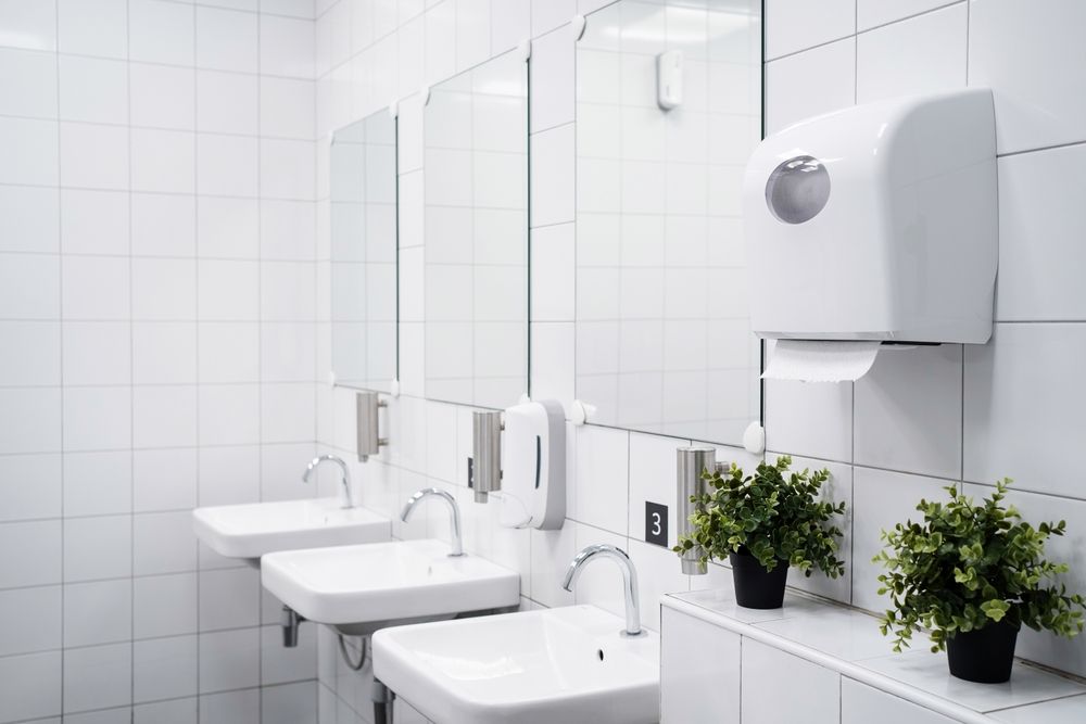 Clean white public restroom with sinks, mirrors, soap dispensers, and potted plants on a tiled counter.
