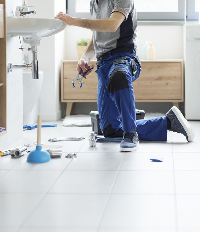 Person kneeling in a bright bathroom, fixing plumbing under a sink with tools scattered on the floor.