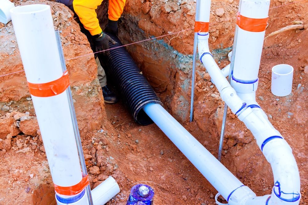 Workers installing white PVC plumbing pipes in a dirt trench with a black corrugated pipe.