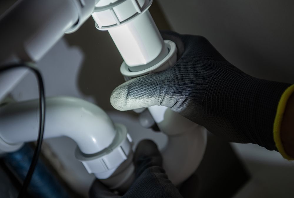 Gloved hands adjusting white PVC pipes under a sink in a dark space