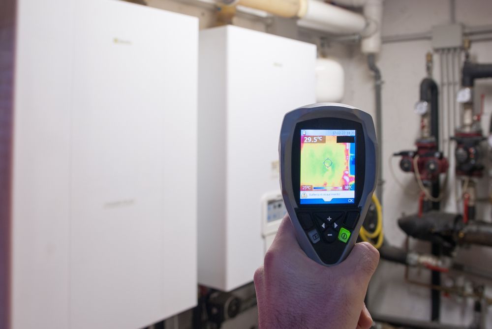Handheld thermal camera aimed at white HVAC units and pipes in a mechanical room