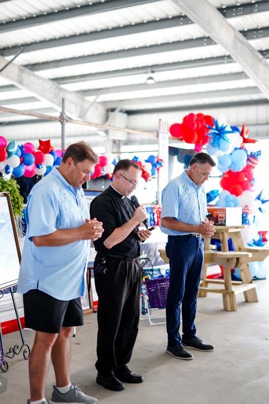 Three people at a decorated indoor event read from papers beside red, white, and blue balloon clusters.