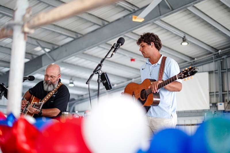 Musicians performing onstage under a metal roof, with red, white, and blue balloons in the foreground