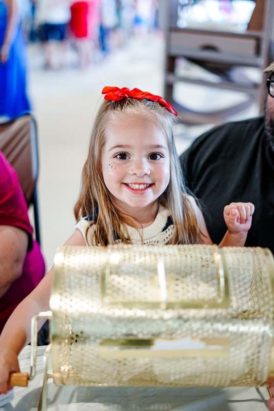 Smiling child beside a golden raffle drum, with a red bow in their hair