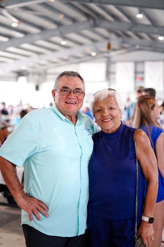 Two smiling adults standing together indoors, one in a light blue shirt and one in a navy sleeveless dress.