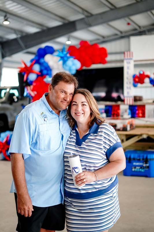 Two people posing together in a decorated indoor event space, holding a drink, with red and blue balloons behind them