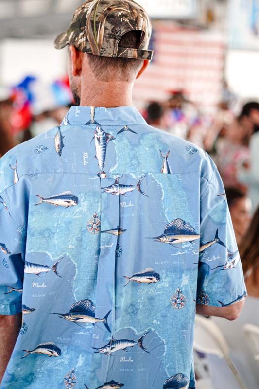 Person in a blue fish-print shirt and camo cap facing away in a busy outdoor crowd