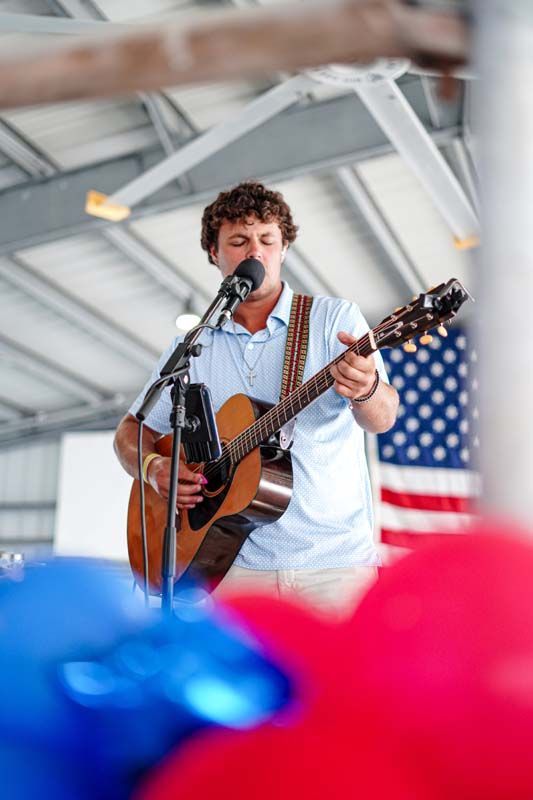 Musician singing and playing acoustic guitar on stage with an American flag backdrop