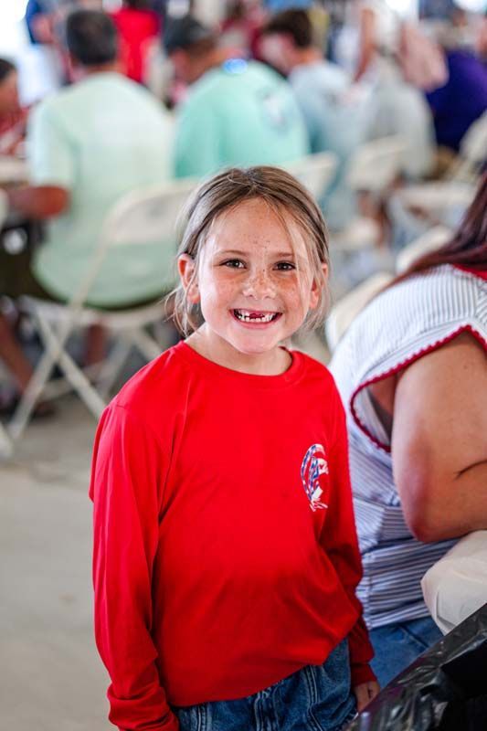 Smiling child in a red shirt standing indoors among seated people