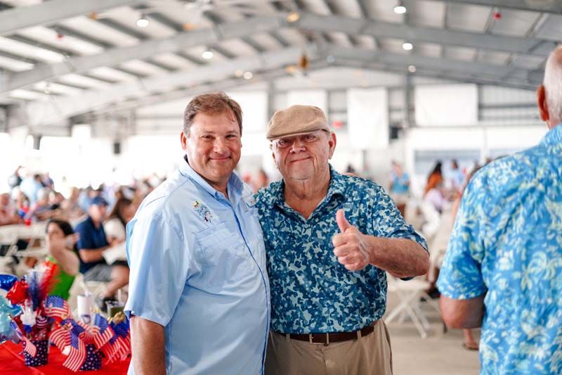 Two men posing at a lively indoor event, one giving a thumbs-up, with colorful tables in the background.