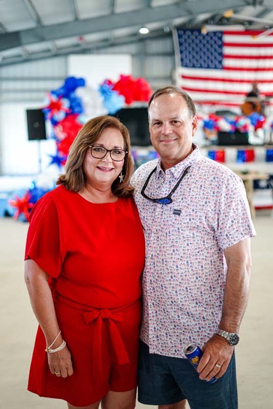 Two people standing indoors in front of an American flag and red, white, and blue decorations.