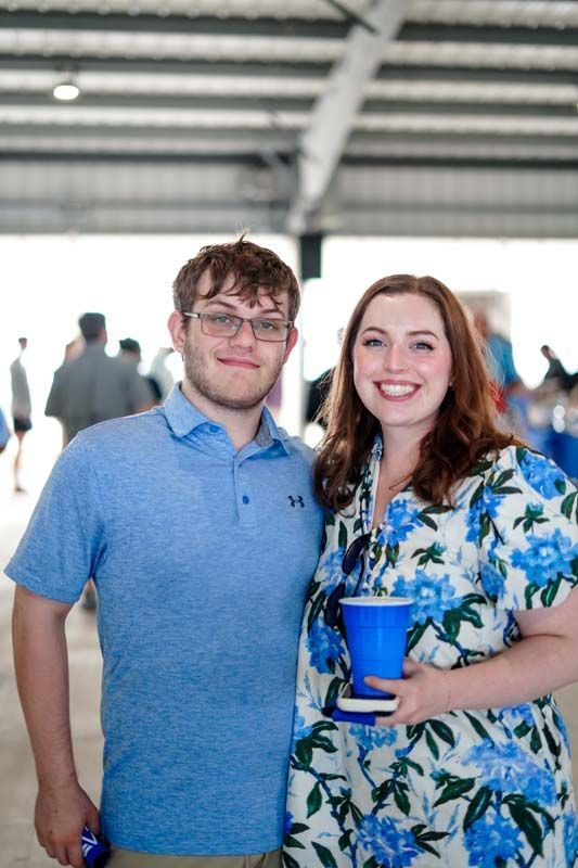 Two people smiling indoors, one holding a blue cup, standing under a metal-roofed pavilion.