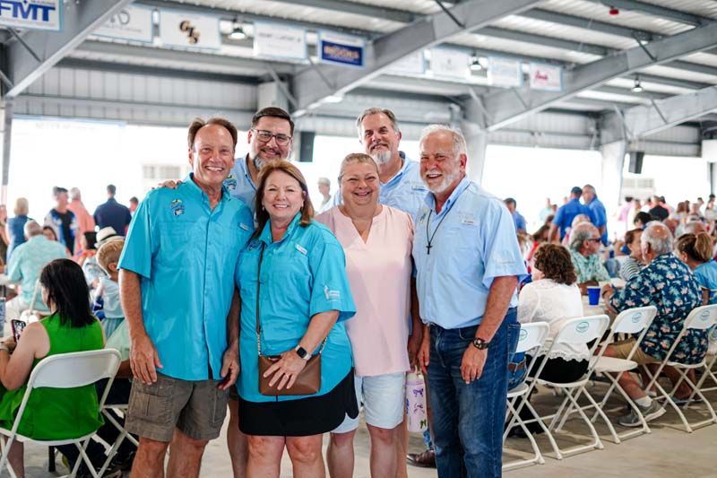 Five smiling people posing together at an indoor event with rows of seated attendees in the background