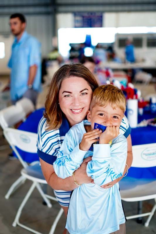 Woman holding a smiling child at a blue-and-white event table, with chairs and people blurred in the background