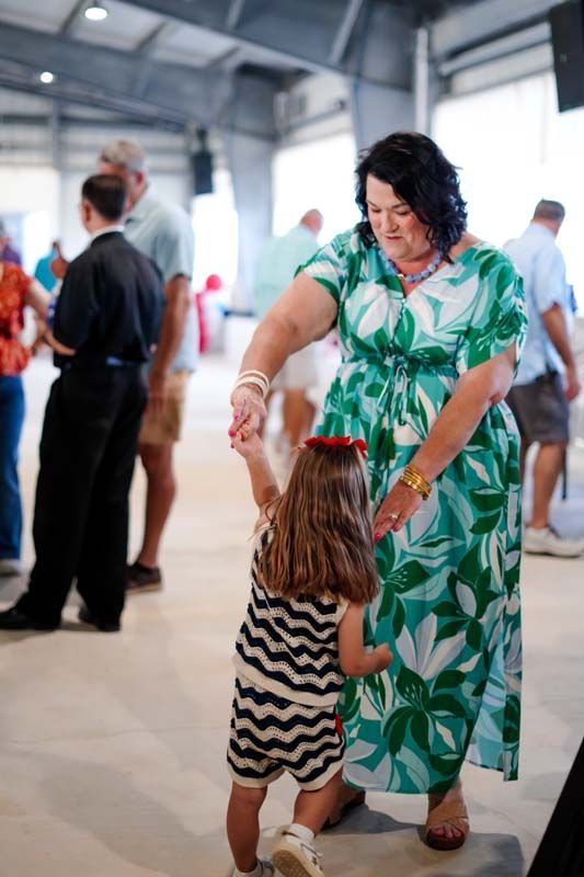 Woman in a green floral dress twirls a child in a striped romper at an indoor event.