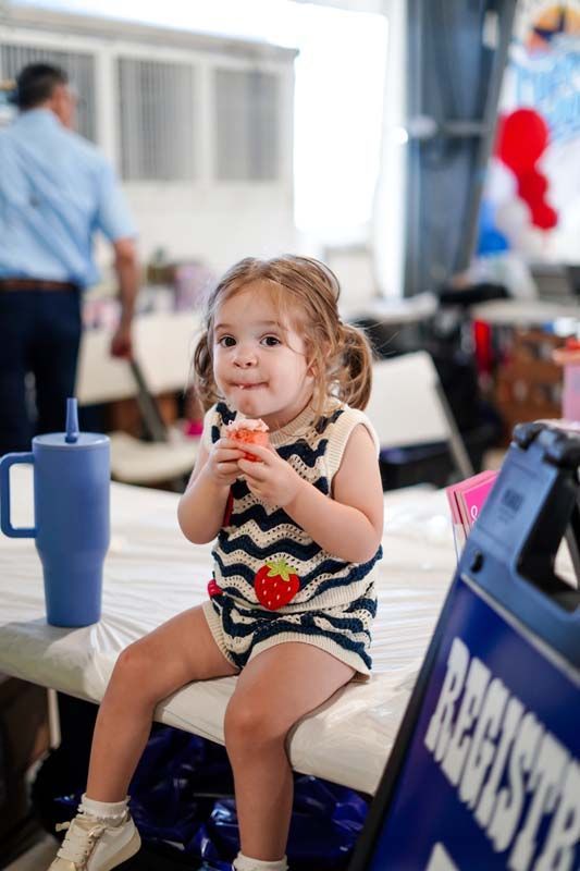 Toddler eating a snack at a table during an indoor event, with a blue cup nearby.