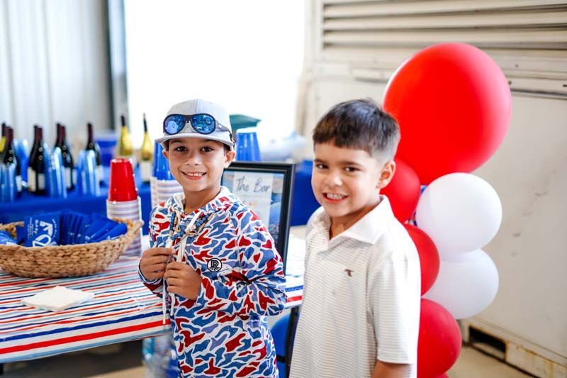Two smiling children beside a festive table with red-and-white balloons and a cake at a party