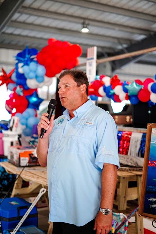 Man speaking into a microphone at a patriotic event with red, white, and blue balloon decorations.