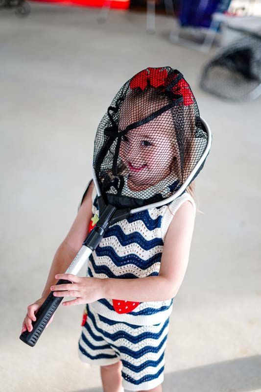 Child holding a tennis racket and smiling behind a black mesh face shield, wearing a blue zigzag outfit.