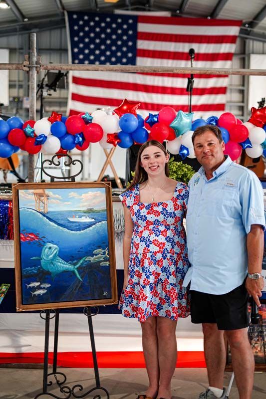 Two people posing beside a framed marine painting, with red, white, and blue balloons and a large American flag behind them.