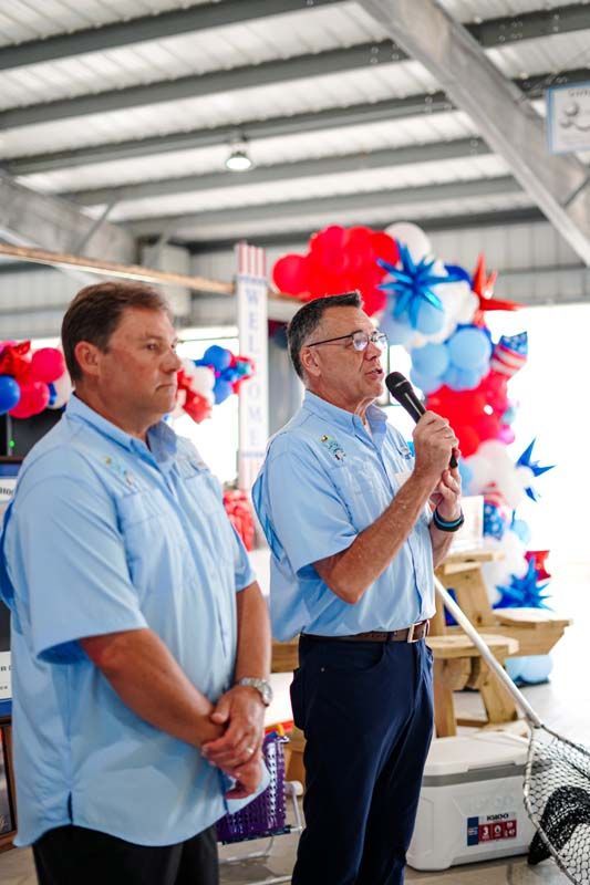 Two men in light blue shirts speak at a decorated event with red, white, and blue balloons.