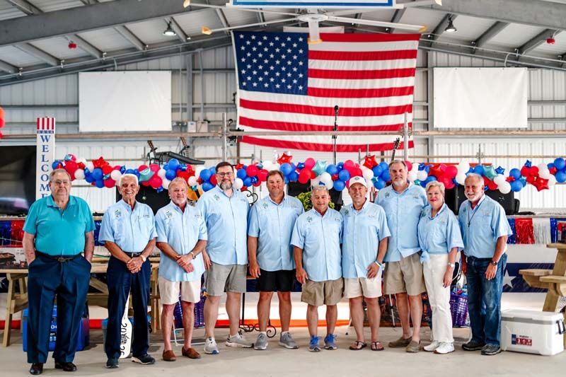 Group of people in blue shirts standing in front of a large American flag and red-white-blue balloons at a factory.