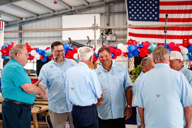 Men in light blue shirts talking at a patriotic indoor gathering with red, white, and blue balloons and a U.S. flag
