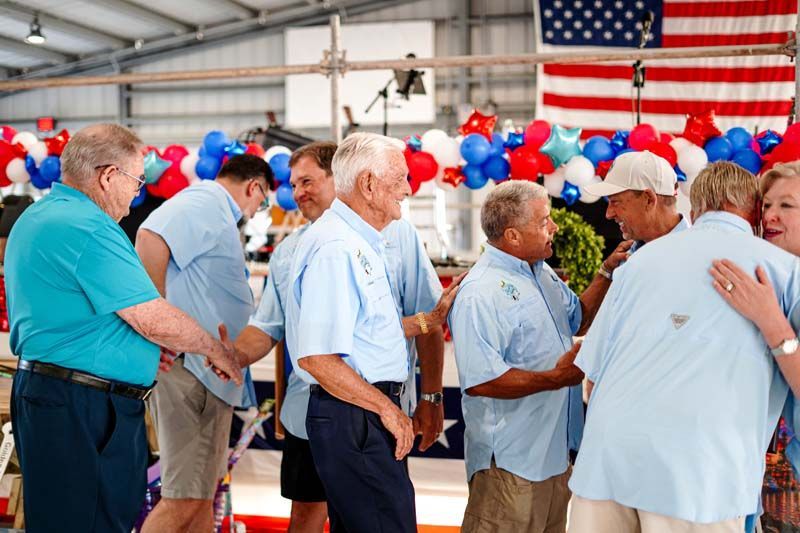 People standing and talking in a hall with red, white, and blue balloons and a large American flag backdrop