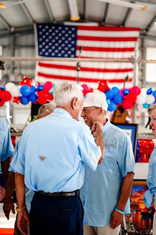 Two men in light blue shirts talking at a patriotic indoor event with American flags and red, white, and blue balloons