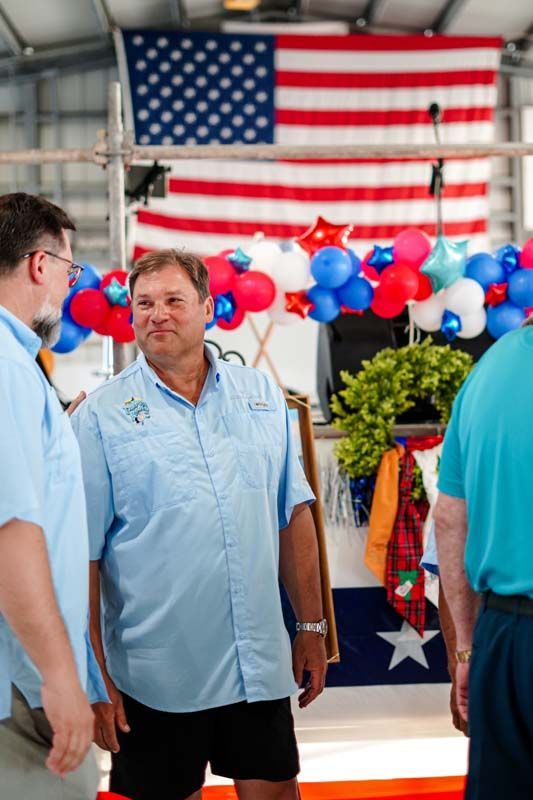 People in blue shirts talking under a large American flag at a festive outdoor booth