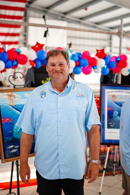 Man in light blue shirt standing at a patriotic event with red, white, and blue balloons and framed displays
