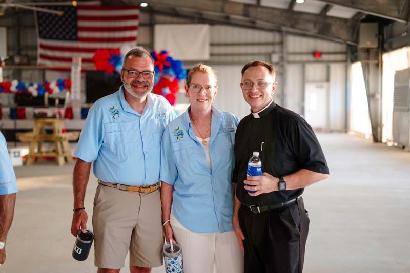 Three people smiling in a hangar, holding drinks, with an American flag in the background.