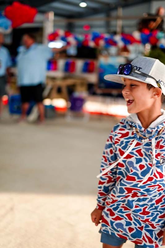 Boy in a red, white, and blue shirt and cap standing outdoors, smiling in a sunny crowd.