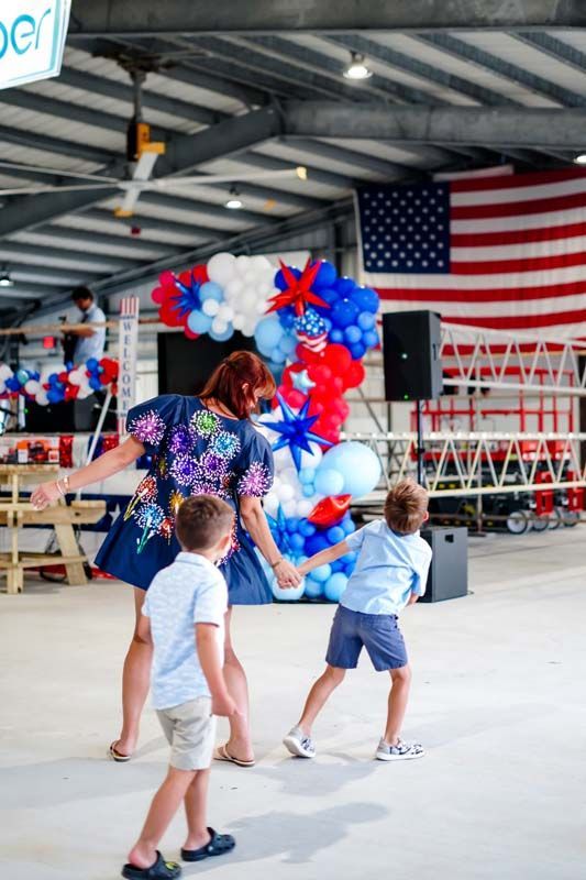 Woman and two children at a patriotic balloon display beneath a large American flag in a covered venue