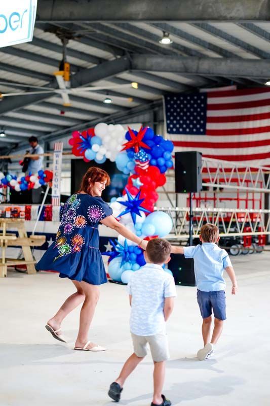 Woman and children in blue outfits dancing at a patriotic event with red, white, and blue balloons and a U.S. flag