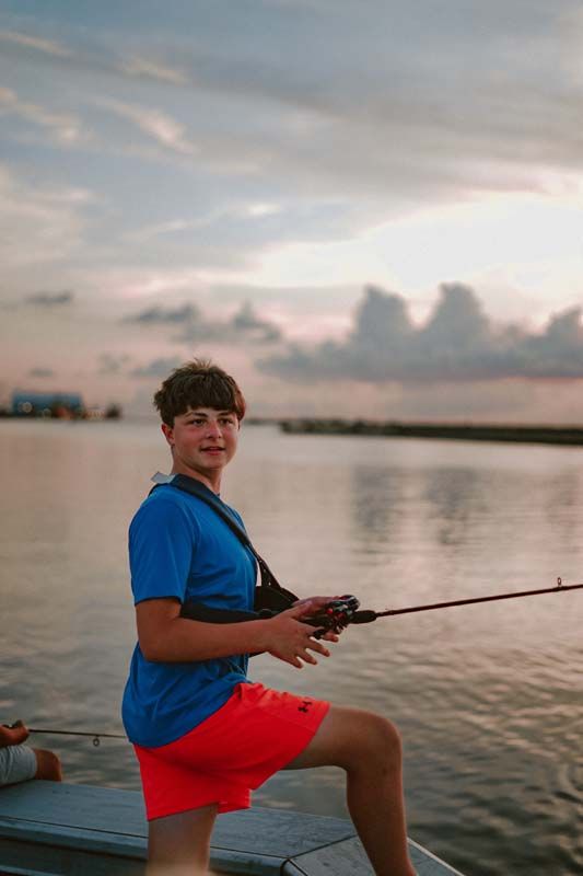 Person fishing from a boat at sunset, wearing a blue shirt and red shorts on calm water.
