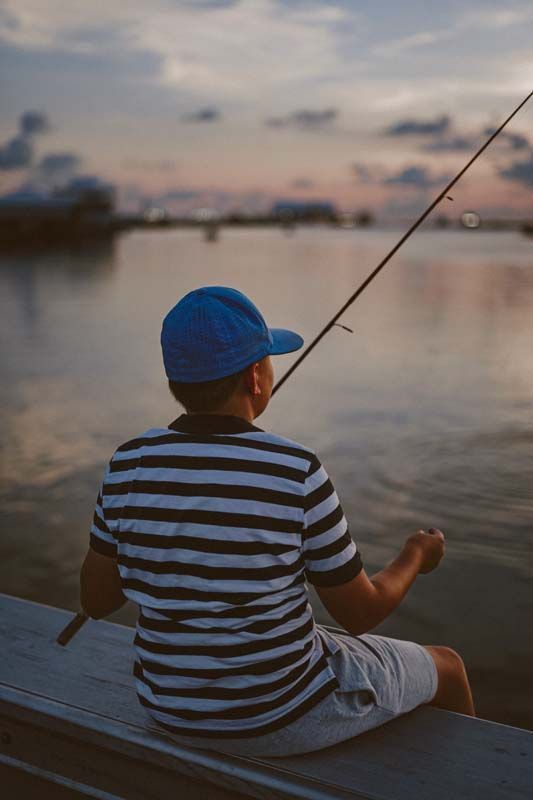 Person fishing at dusk from a dock, wearing a blue cap and striped shirt, with calm water and a pink sky