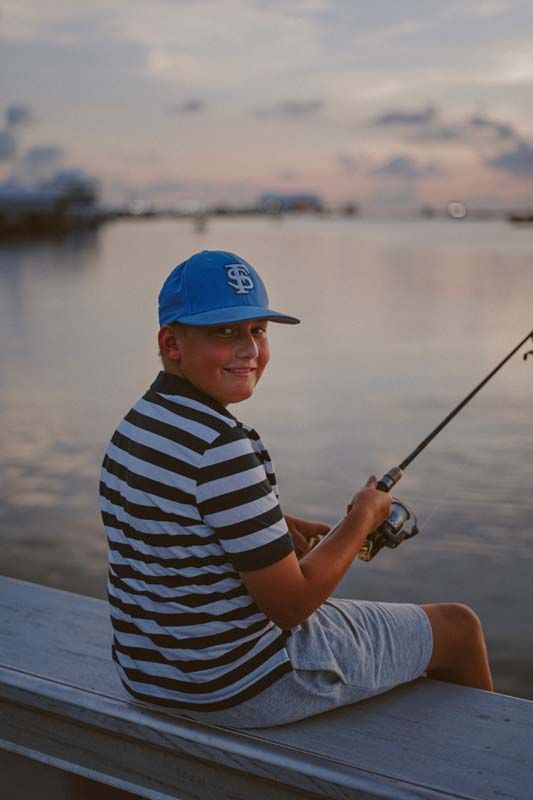 Boy in blue cap and striped shirt fishing from a dock at sunset.