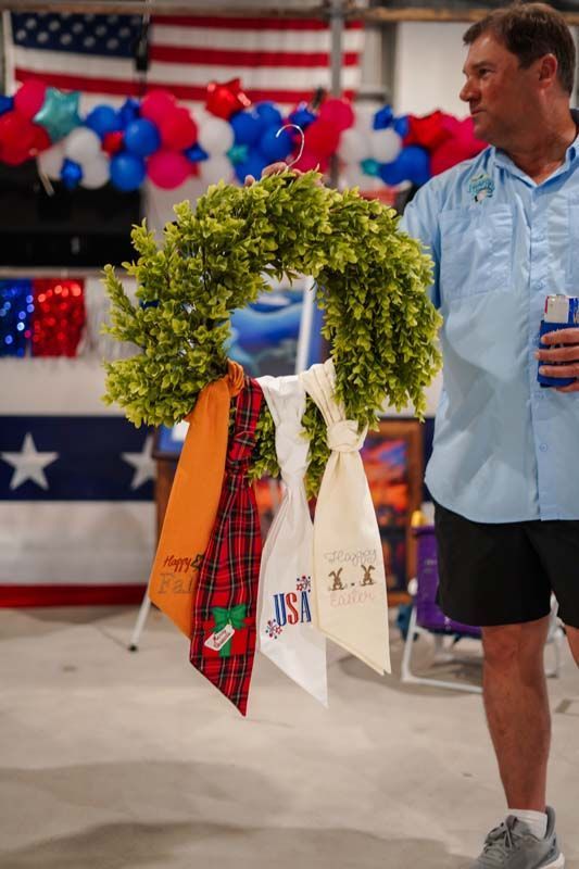 Decorated green wreath with ribbons and tags held beside a man at a patriotic indoor event