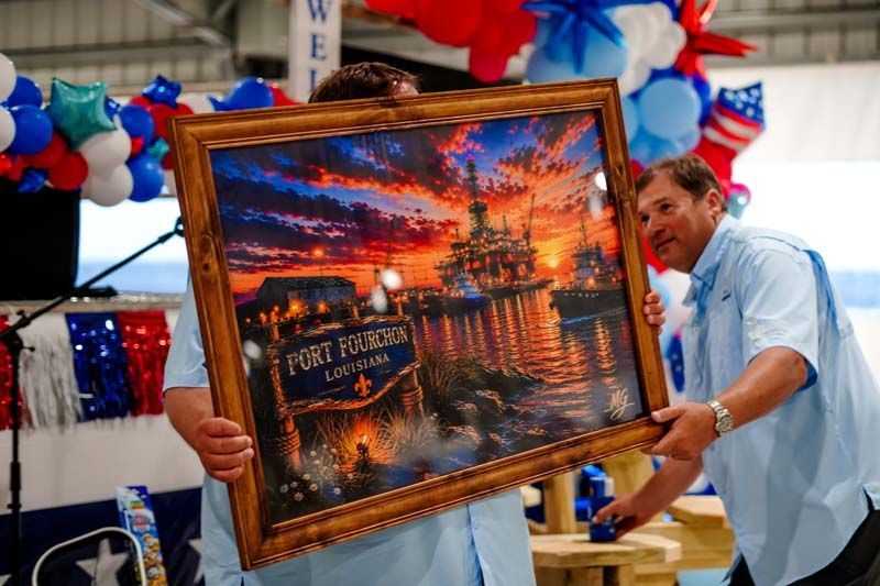 Two men hold a framed sunset harbor painting at an art fair, with patriotic decorations behind them