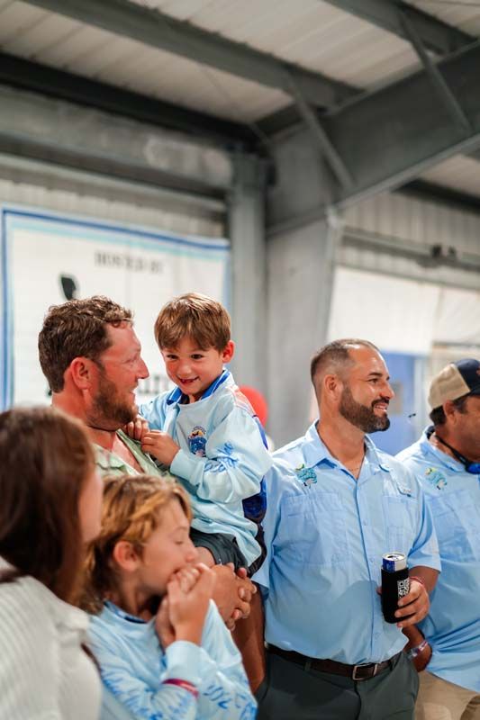 People gathered indoors at an industrial event, one man holding a smiling child in a blue jacket.