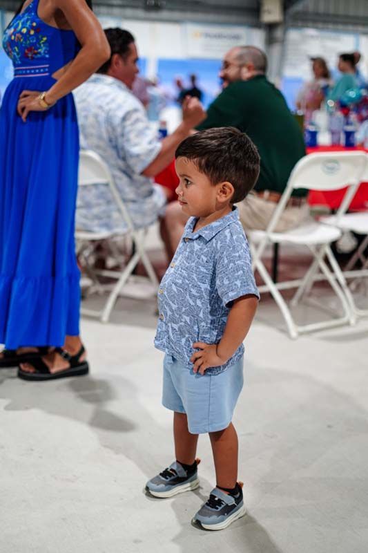 Child standing in a blue shirt and shorts at an outdoor gathering with white folding chairs and people nearby